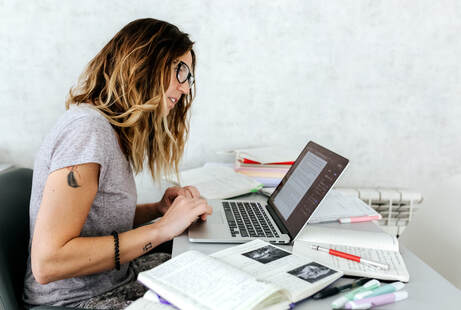 Young Adult Caucasian Female working on laptop at desk in home office, wearing glasses and gray t-shirt, with tattoos on R arm |  Creativity Counseling for Creative People in Maryland and Virginia, Creativity Counseling Online for Creative Adults and Children with ADHD