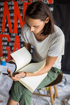 Young person seated on stool wearing white t-shirt and green trousers, holding sketchbook | ADHD, Creative People, Perfectionism and Strong Emotions are related features; find relief with Creativity Counseling available online Maryland and Virginia at Creatively, LLC