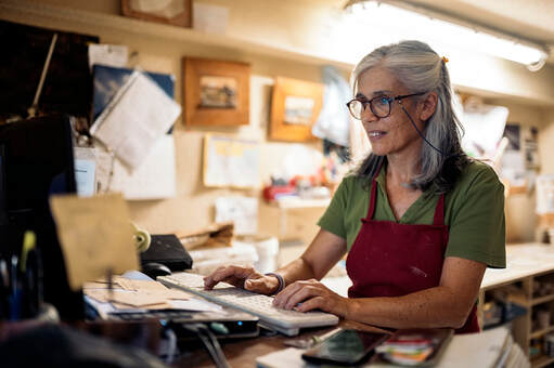 Adult 50+ female working in art studio at computer keyboard, wearing green shirt, red apron and glasses | Online Counseling for Creatives, Virtual Therapy for Creative People With Cindy Cisneros, LCPC, LPC, Creativity Coach at Creatively, LLC