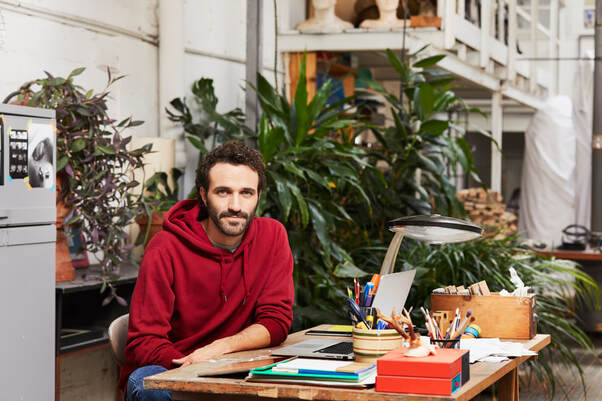 Adult Caucasian male in red sweatshirt seated at table with art supplies and laptop in apartment with plants in background | Creativity Coaching benefits Artists, Writers, Musicians, Actors, Content Creators and more, available online in MD, VA, ME, NY, GA, UT, CA and beyond throughout the USA, Canada and the UK