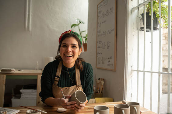 Latina in brown apron, green shirt, and scarf, in studio with window during day at Left, smiling |  Uncover your Creative Personality at Creatively, LLC WITH CINDY Cisneros, Creativity Coaching and Creativity Coaching, Online
