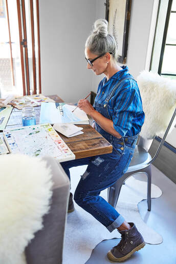 Over 50 Caucasian female painting at studio desk wearing blue shirt and overalls | Online Creativity Coaching for Artists, Writers and Performers with Cindy Cisneros, Online Creativity Coach in Maryland, Virginia, Utah, Maine, Georgia, New York, California and throughout the USA, UK and Canada