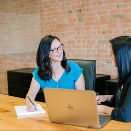 Latina women seated with laptop at conference table, smiling | Person Centered Therapy with Hybrid Creativity Coaching for Creative People and Creative Professionals for emotional and career support