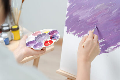 Picture of a person's hands holding a palette and painting with purple paint. Working artists can get creative support with creativity coaches in Germany, the United States, Canada, the UK and beyond here.