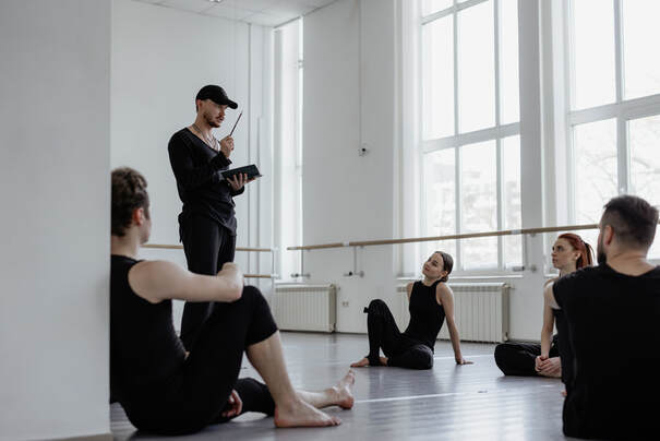 Dancers wearing black, seated on floor, around instructor with tablet, in dance studio |  Therapy for Creatives in Maryland and Creativity Counseling in Maryland for Gaslighting