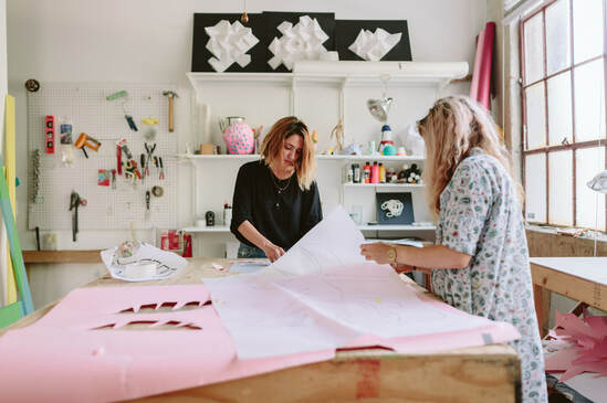 two women at work table in art studio with large sheets of paper, having conversation | Join the Virtual Artists Residency at Creatively LLC and connect with other Artists to grow your Creative Practice