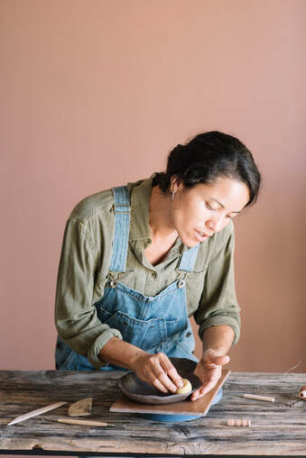 Latin American Female wearing overalls and green shirt hand building with clay at wooden table |  Start Creativity Coaching Today! Online Creativity Coaching for Artists With Cindy Cisneros Nationwide including GA, MD, VA, ME, UT, NY, CA and beyond!