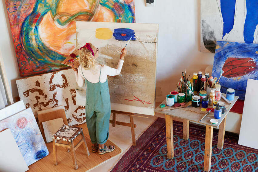 Caucasian women, blonde, in green coveralls, facing easel, standing, painting, with canvases and paints | Therapy for Creatives and Online Coaching for Creatives, Creative Personalities Supported at Creatively, LLC