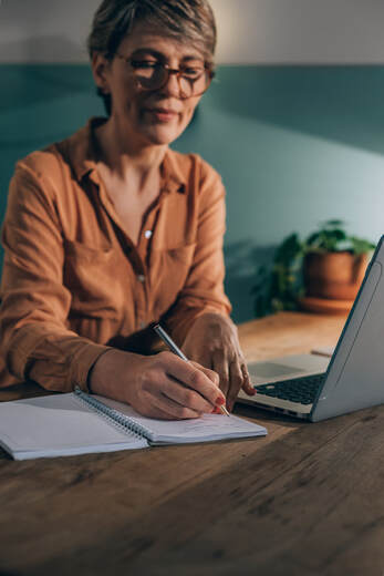 50+ female at desk with notebook and laptop, wearing orange shirt and glasses | Concierge Counseling Services Online in Maryland With Cindy Cisneros, LCPC-S, LPC at Creatively, LLC