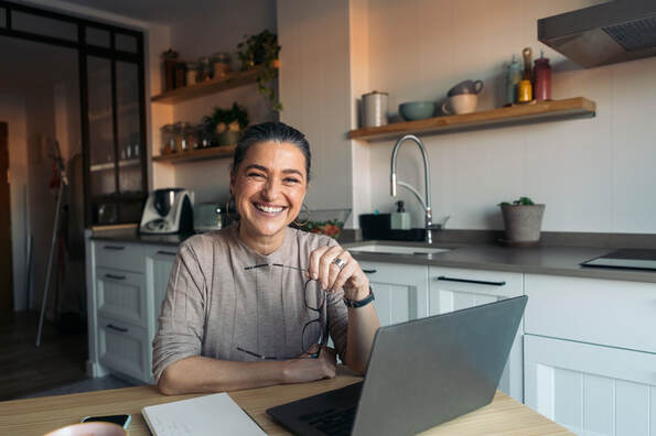 Adult female seated in kitchen with laptop, facing camera and smiling broadly | Relief from ADHD for Creative People at Creatively, LLC with Creativity Counseling, Available Online in Maryland and Virginia