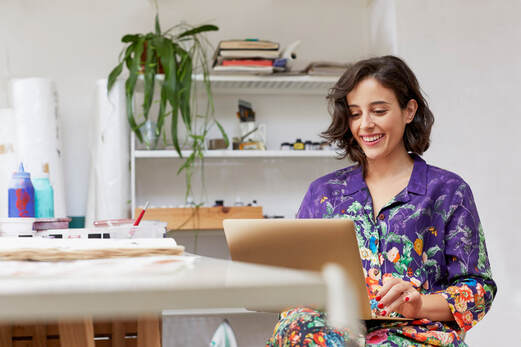 Adult Caucasian female wearing purple floral blouse in studio seated at desk working at laptop, smiling |  Creativity Counseling for Stress and Anxiety for Creative People in Maryland and Virginia 