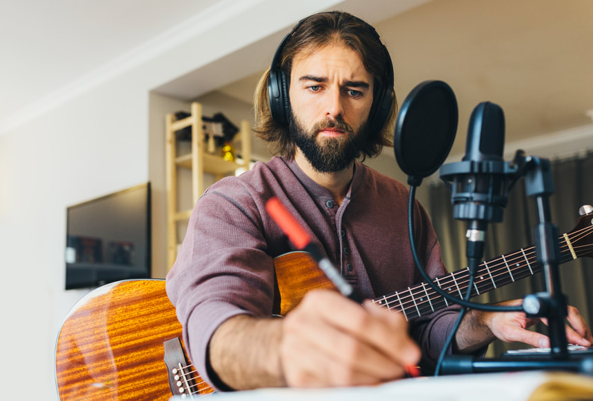 Caucasian male with long hair and beard in home recording studio holding guitar, at keyboard, making notes with red pen |  Online Creativity Coaching for Musicians in Utah: Creativity Coaching in MD, GA, ME, VA, UT and beyond for Musicians
