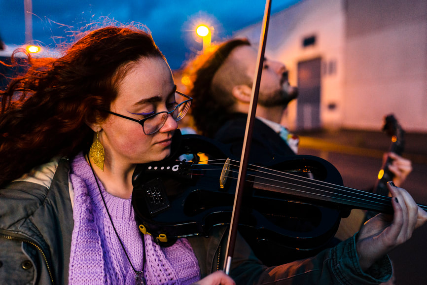 female with red hair and glasses playing violin with male bandmate in beard playing stringed instrument in background outside in industrial area at night |  Online Creativity Coaching With Cindy Cisneros for Musicians: Creativity Coaching in Maryland and Virginia and beyond