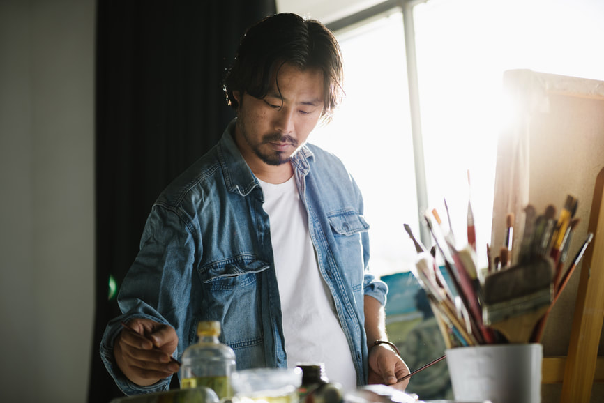 Adult Asian Male painting at desk in front of window wearing denim shirt | Creativity Coaching for Creative Blocks: Online Creativity Coaching for Artists in MD, VA, NY, UT, GA, ME, CA and beyond