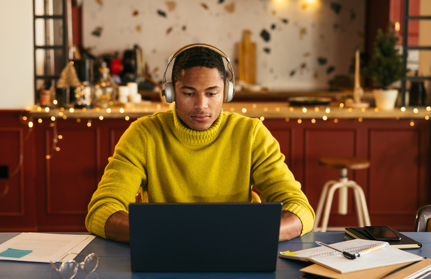 young Latino man in yellow sweater and headphones writing at laptop with papers on table | Creativity Coaching available: Online Coaching for Creatives serving Writers, Performers, Artists and Entrepreneurs, Online Creativity Coaching in MD, VA, GA, UT, CA, NY, ME and beyond