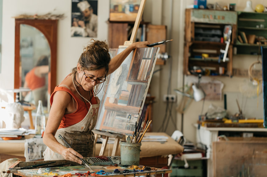 Adult 50+ female wearing apron, red shirt and glasses in painting studio painting at easel |  Creativity Counseling to Live your Authentic Live and Create with Ease for Creative Artists in Maryland and Virginia